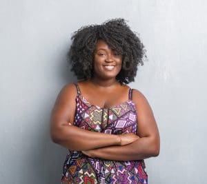 Young african american woman over grey grunge wall wearing colorful dress happy face smiling with crossed arms looking at the camera. Positive person.